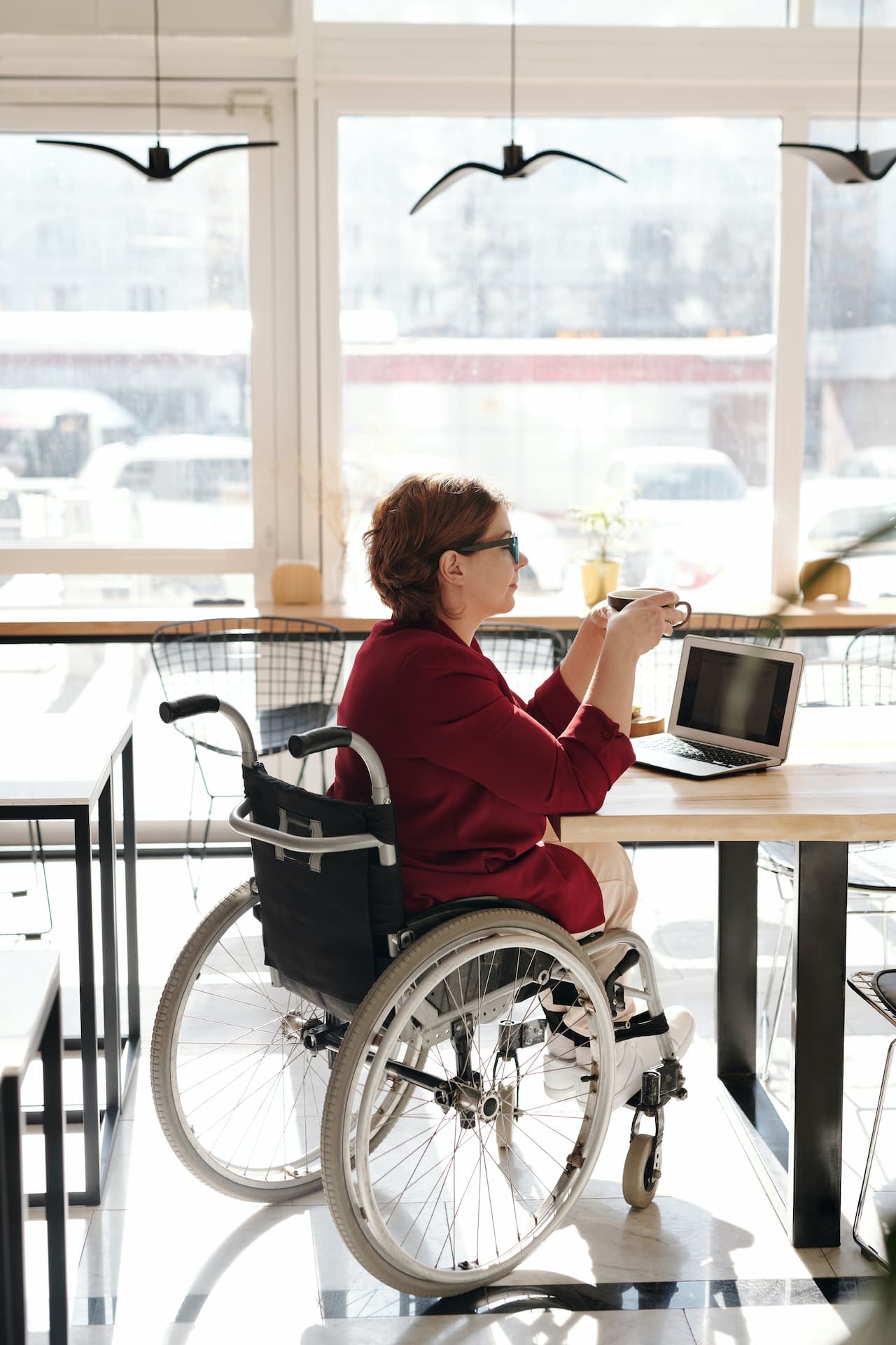 Woman in wheelchair using laptop, drinking coffee in cafe.